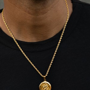May include: A gold rope chain necklace with a gold pendant featuring a depiction of Saint Christopher. The pendant has the words "Saint Christopher Protect Us" inscribed around the edge.