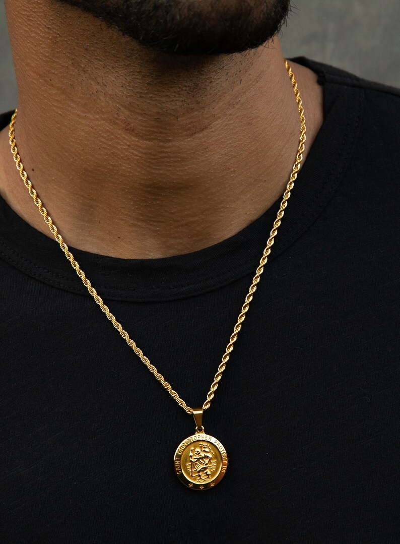 May include: A gold rope chain necklace with a gold pendant featuring a depiction of Saint Christopher. The pendant has the words "Saint Christopher Protect Us" inscribed around the edge.