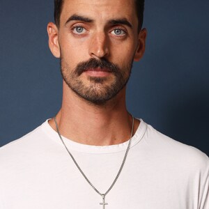 May include: A man with a mustache wearing a white t-shirt and a silver chain necklace with a small silver cross pendant.