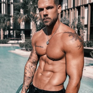 May include: A shirtless man with tattoos on his arms and chest stands by a pool. He is wearing black jeans and a silver chain necklace with a pendant. The text "vivre en la lluvia" is tattooed on his chest.