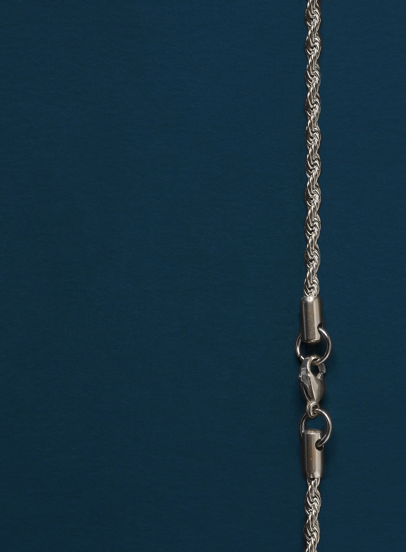 May include: Silver-colored rope chain necklace with a lobster clasp, displayed against a dark teal background. The chain has a twisted rope design, and the clasp is visible at the bottom.
