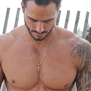 May include: A man wearing a gold chain necklace with a small gold cross pendant. The chain is 22 inches long. The man has short dark hair and a beard. He is wearing a white tank top and has a tattoo on his right arm. The text "MIKE IS WEARING THE SMALL CROSS AT 22 INCHES" is visible at the bottom of the image.