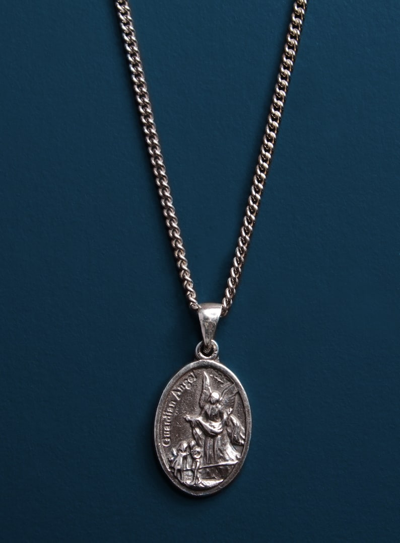 Pu&ograve; includere: Collana con catena d'argento con un ciondolo ovale d'argento con un designo di angelo custode. Il ciondolo ha le parole "Guardian Angel" incise su di esso.