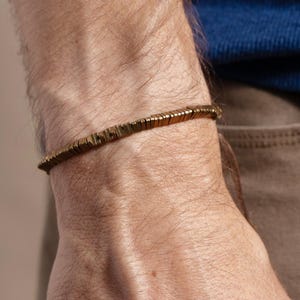 May include: A gold-colored bracelet composed of small, rectangular links. The bracelet is worn on a wrist, with a blue shirt and brown pants visible in the background. The bracelet appears to be made of metal.