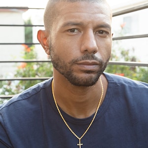 May include: A man with short, dark hair and a beard is wearing a dark blue t-shirt and a gold chain with a cross pendant.