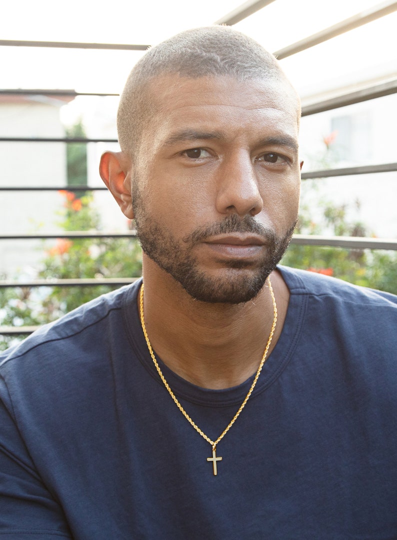 May include: A man with short, dark hair and a beard is wearing a dark blue t-shirt and a gold chain with a cross pendant.