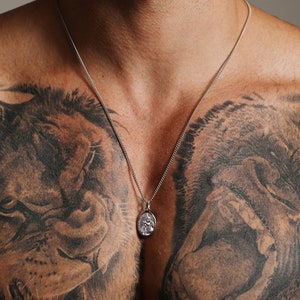 Pu&ograve; includere: Una collana a catena d'argento con un piccolo ciondolo ovale con un designo dettagliato. La collana &egrave; indossata da una persona con tatuaggi sul petto.