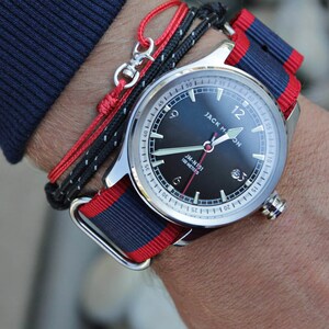 May include: A silver wristwatch with a black face and red and blue striped nylon band. The watch face has white markings and the text "JACK HON" and "JM-N191 100 METERS".