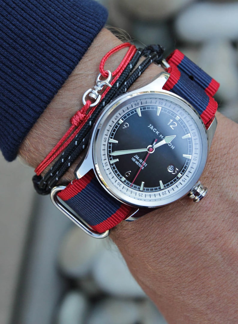 May include: A silver wristwatch with a black face and red and blue striped nylon band. The watch face has white markings and the text "JACK HON" and "JM-N191 100 METERS".