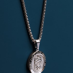 May include: A silver chain necklace with a round pendant featuring a depiction of Saint Christopher. The pendant has the words "Saint Christopher Protect Us" inscribed around the edge.