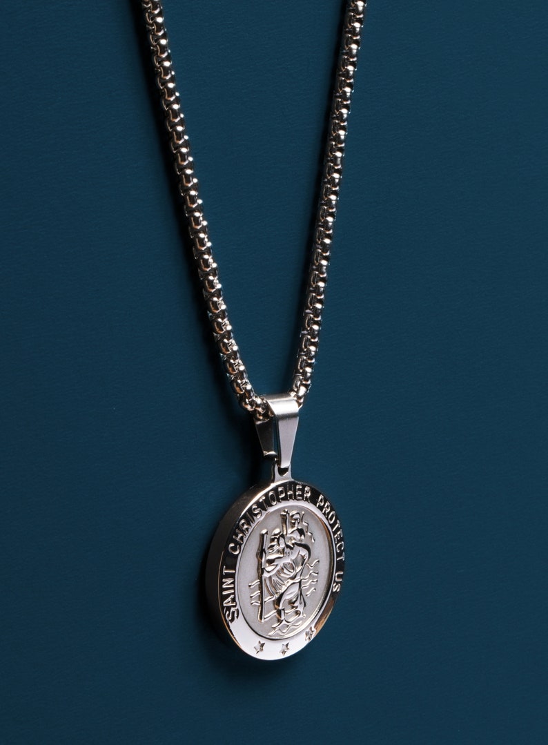 May include: A silver chain necklace with a round pendant featuring a depiction of Saint Christopher. The pendant has the words "Saint Christopher Protect Us" inscribed around the edge.