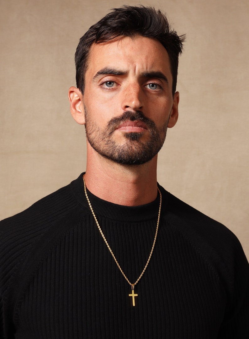 May include: A gold-colored cross pendant hangs from a gold chain. The cross is a simple, classic design. The chain is worn over a black ribbed sweater.
