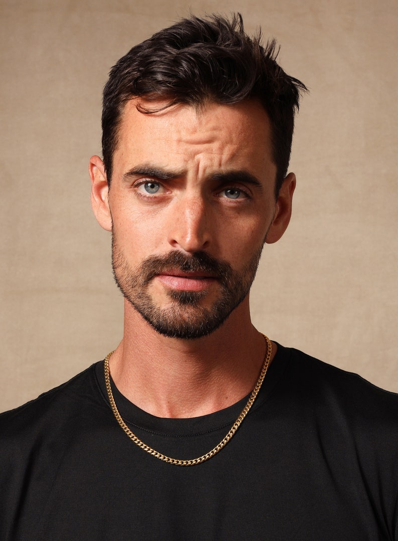 May include: A man with short, dark hair and a beard is wearing a black t-shirt and a gold chain necklace.