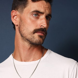 May include: A man with short dark hair and a beard is wearing a white t-shirt and a silver chain necklace with a cross pendant.