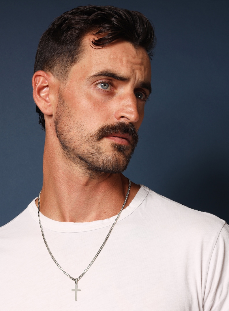 May include: A man with short dark hair and a beard is wearing a white t-shirt and a silver chain necklace with a cross pendant.