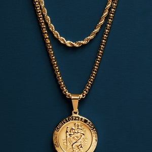 May include: A gold chain necklace with a gold pendant featuring a depiction of Saint Christopher. The pendant reads "Saint Christopher Protect Us".