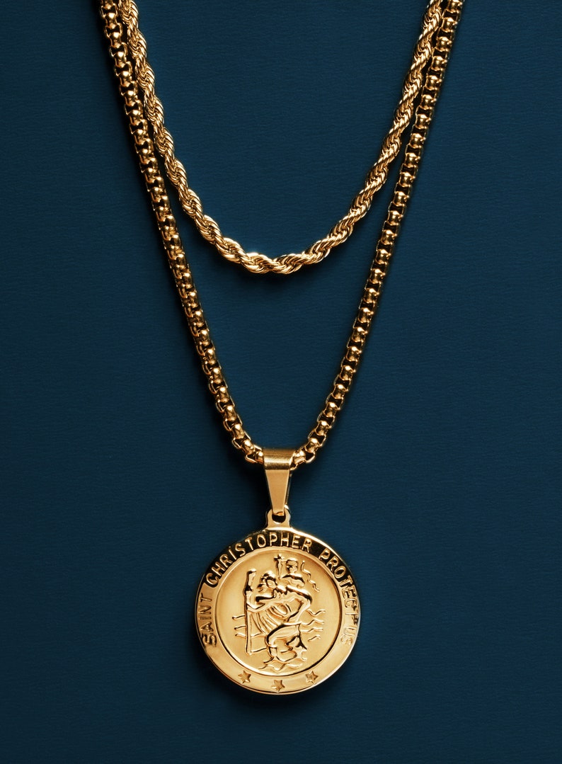 May include: A gold chain necklace with a gold pendant featuring a depiction of Saint Christopher. The pendant reads "Saint Christopher Protect Us".