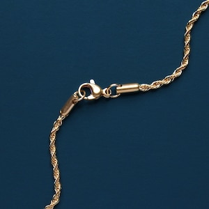 May include: Close-up of a gold-colored rope chain necklace with a lobster clasp. The chain is made of twisted strands, creating a textured appearance. The background is a solid, dark blue color.