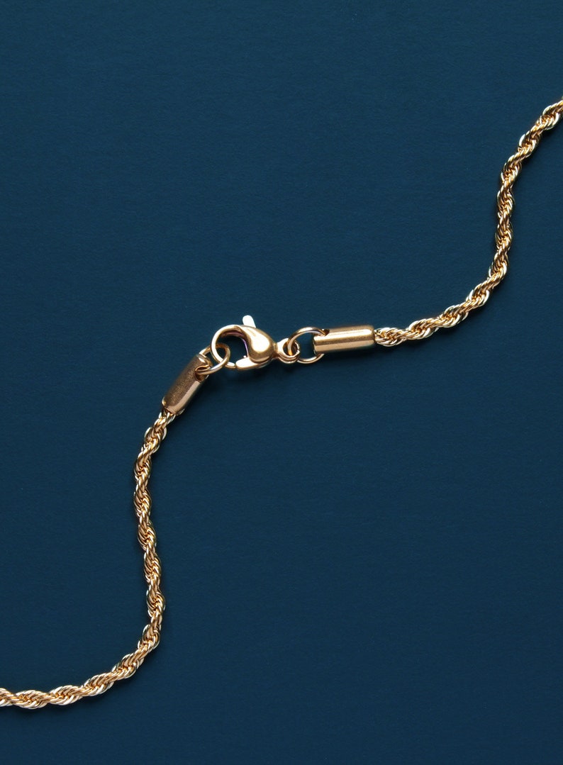 May include: Close-up of a gold-colored rope chain necklace with a lobster clasp. The chain is made of twisted strands, creating a textured appearance. The background is a solid, dark blue color.