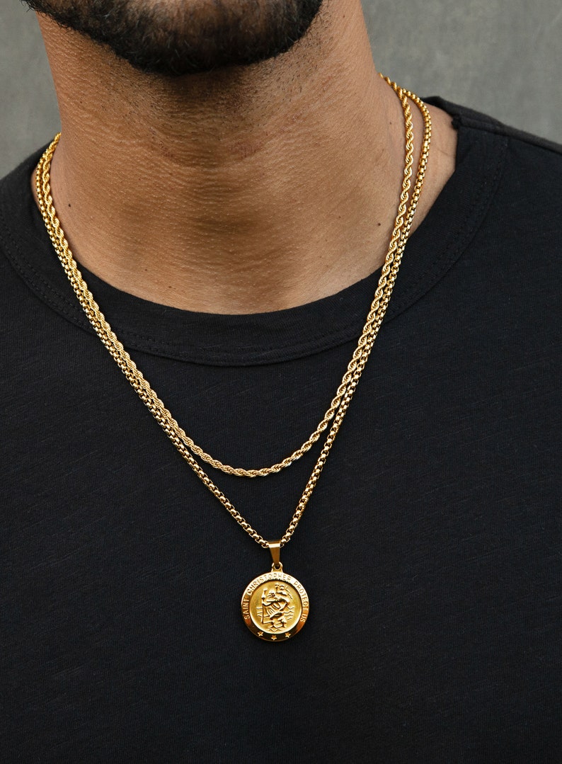 May include: A gold chain necklace with a gold pendant featuring a Saint Christopher medal. The pendant has the words "Saint Christopher Protect Us" inscribed around the edge.