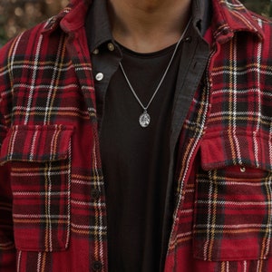 Pu&ograve; includere: Un uomo indossa una camicia di flanella a quadri rossi e neri sopra una maglietta nera. Indossa una collana a catena d'argento con un piccolo ciondolo.
