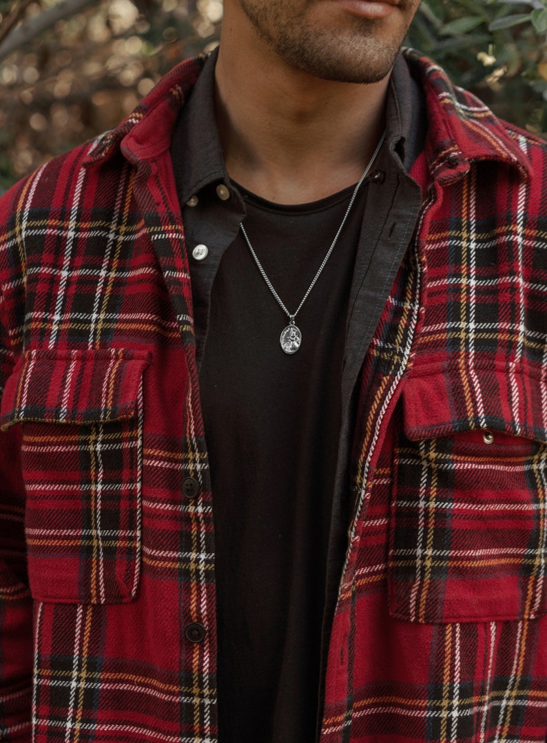 Pu&ograve; includere: Un uomo indossa una camicia di flanella a quadri rossi e neri sopra una maglietta nera. Indossa una collana a catena d'argento con un piccolo ciondolo.