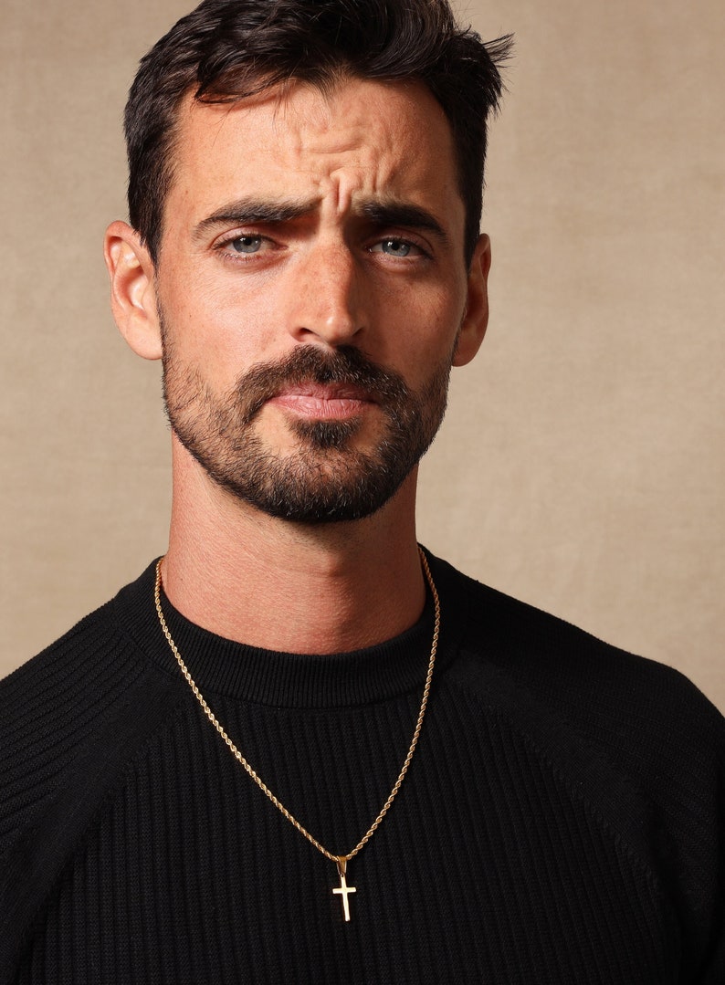 May include: A man wearing a black sweater with a gold rope chain necklace and a small gold cross pendant.