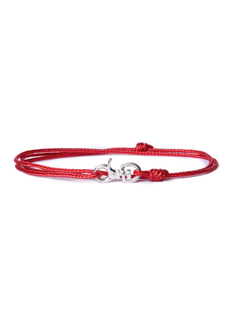 May include: A red cord bracelet with a silver clasp. The bracelet is made of a thin, braided cord and has a simple, elegant design.