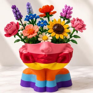 May include: A multi-coloured planter shaped like a human head resting on hands, featuring horizontal stripes in pink, red, yellow, orange, light blue, and purple. The planter is filled with a variety of colourful plastic brick-built flowers.
