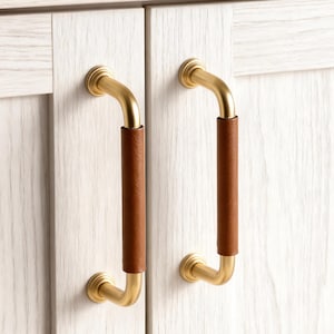 Puede incluir: Dos tiradores de puerta de armario con acabado de latón y agarre de cuero marrón. Los tiradores están montados en un armario blanco con una textura de veta de madera clara. Un tarro de cristal con cristales amarillos está encima del armario.