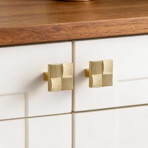 May include: Two square, gold-coloured cabinet knobs with a textured, grid-like pattern are mounted on a white cabinet. A wooden shelf with a brown finish sits above the cabinet. A wooden diffuser with reeds is on the shelf.
