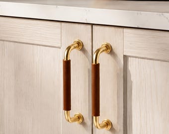 Può includere: Due maniglie per armadietto con struttura in metallo color oro lucido e un rivestimento in pelle marrone scuro sull'impugnatura centrale. Le maniglie sono montate verticalmente su ante di armadietto in legno chiaro con venature.