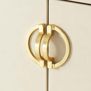 May include: Close-up of two cabinet doors with gold-colored hardware. The hardware features a circular design with a central bar and a curved handle. The cabinet doors are a light cream color, providing a neutral backdrop for the hardware.