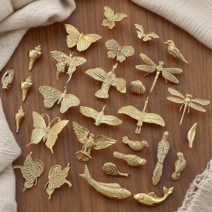 Peut inclure: Une collection de petites figurines métalliques dorées disposées sur une surface en bois. L'assortiment comprend des papillons, des oiseaux, des libellules, des poissons et d'autres formes inspirées de la nature. Les figurines sont détaillées et semblent être des objets décoratifs.