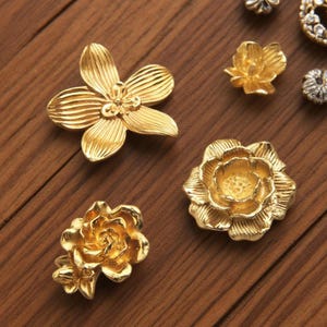 May include: A collection of gold-colored floral embellishments, including a four-petal flower, a small multi-petal flower, and a rose-like design. The pieces are arranged on a wooden surface, showcasing their intricate details.