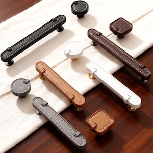 Könnte beinhalten: Eine Auswahl an Schrankgriffen und -knöpfen aus Leder in Schwarz, Braun, Weiß und Grau. Die Griffe sind rechteckig mit Kontrastnähten, die Knöpfe rund oder quadratisch. Präsentiert auf einem weißen Tuch und einer Holzoberfläche.