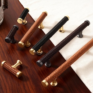 May include: A collection of cabinet hardware featuring woven leather handles in various colors, including black, brown, and tan. The handles have gold or dark metal accents and are displayed on a wooden surface.