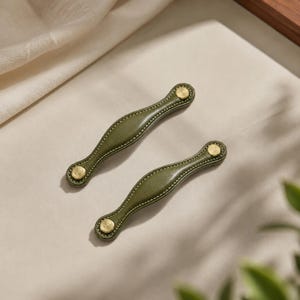 May include: Two olive green leather drawer pulls with a curved, elongated shape. Each handle features light-colored stitching along the edges and is secured at both ends by circular, brushed brass-toned metal fasteners with a central screw detail.