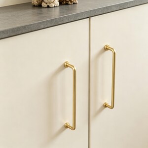 May include: Two vertical, brushed gold-toned metal cabinet handles with a cylindrical bar design and circular mounting bases, attached to smooth, off-white cabinet doors. The handles feature a simple, clean, and functional hardware structure.
