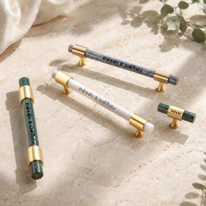 May include: A collection of cabinet hardware featuring cylindrical bars and knobs with gold-toned metal mounts. The pieces include a dark grey marble bar, a white bar, a green marble bar, and a small green marble knob, all printed with Peter & Wendy.