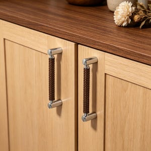 May include: Two vertical cabinet handles featuring a dark brown braided leather grip section secured by polished silver-toned metal end caps. The handles are mounted onto light-toned wooden cabinet doors with a visible vertical grain pattern.