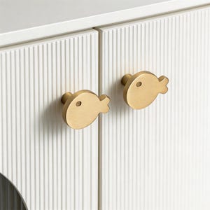 May include: Two gold-colored, fish-shaped cabinet knobs are mounted on a white cabinet with vertical ribbed detailing. The knobs have a simple, rounded design with a small circular eye detail. The cabinet doors are closed.