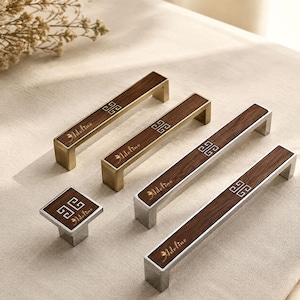 May include: A collection of four cabinet hardware pieces featuring a dark wood inlay with a gold or silver metal frame. Each piece displays the word Adeline in script and a geometric square pattern. The set includes three rectangular pulls and one square knob.