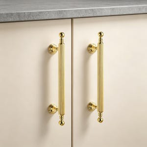 May include: Two vertical cabinet pulls made of polished gold-tone metal. Each handle features a long, cylindrical, fluted center section with rounded, decorative finials at the top and bottom, attached to the cabinet doors by two circular mounts.