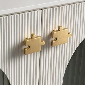 May include: Close-up of a white cabinet with vertical ribbed detailing and two gold-colored puzzle piece shaped cabinet pulls. The cabinet doors are closed, and the pulls are centered on each door.