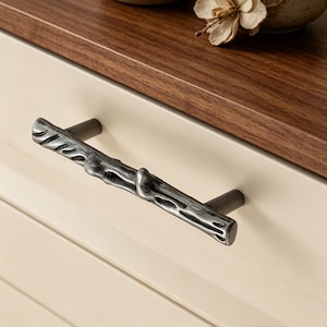 Könnte beinhalten: Ein horizontaler Schubladengriff mit einer texturierten, organischen, astähnlichen Optik in einem dunklen, verwitterten Metallfinish. Der Griff ist an einer glatten, cremefarbenen Schubladenfront montiert und verfügt über zwei zylindrische Befestigungspunkte.