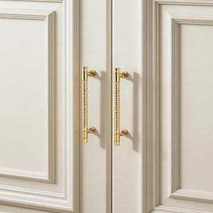 Puede incluir: Dos tiradores dorados para armario con un diseño vertical texturizado. Los tiradores están montados en una puerta de armario blanca. El herraje tiene un estilo clásico y elegante.