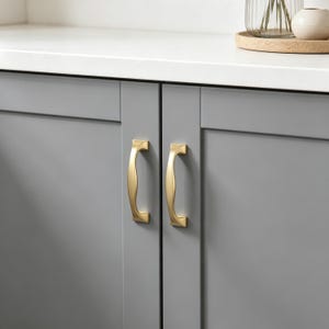 May include: Two brushed gold cabinet pulls with a curved, ergonomic design mounted vertically on two adjacent gray cabinet doors. The hardware features a smooth, metallic finish and a solid construction, providing a functional grip for opening.