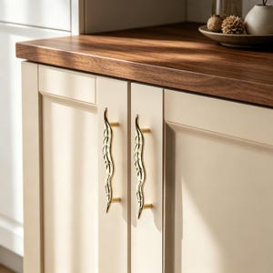May include: Two vertical cabinet handles in a polished gold finish, featuring a slender, wavy design that mimics the shape of a leaf with etched vein details. These metallic pulls are mounted onto cream-colored cabinet doors with a flat panel frame.