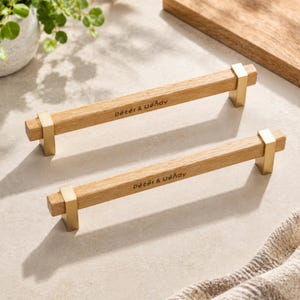 Puede incluir: Dos tiradores lineales para muebles compuestos por una barra de madera clara natural sujeta por dos soportes de metal dorado cepillado. Cada mango de madera tiene grabado el texto Peter & Wendy en una fuente oscura, diseño horizontal.